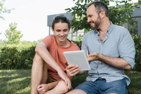 Padre barbudo y feliz hijo adolescente mirando tableta digital mientras está sentado en el parque verde - foto de stock