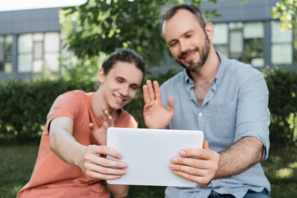 Alegre papá y adolescente hijo agitando las manos mientras tiene videollamada en la tableta digital - foto de stock