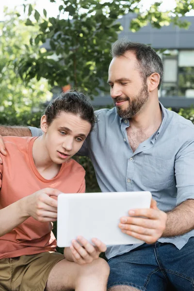Бородатий тато дивиться на сина-підлітка, використовуючи цифровий планшет у зеленому парку — стокове фото