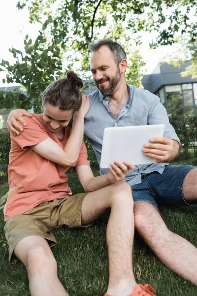 Радісний тато дивиться на підлітка сина і цифровий планшет, сидячи в зеленому парку — стокове фото