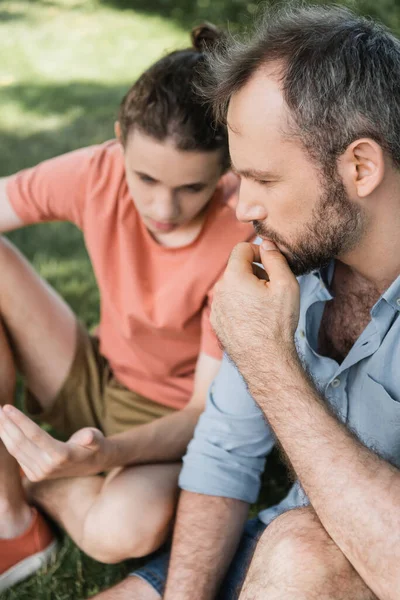 Высокий угол зрения застенчивого и босоногого отца, сидящего рядом с сыном-подростком в зеленом парке — стоковое фото
