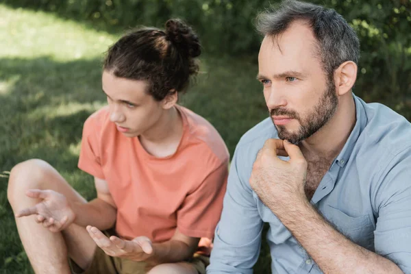 Pensive and bearded father sitting next to teenage son while having conversation in green park — Stock Photo