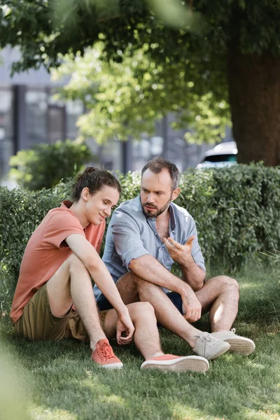 Barbudo padre hablando con hijo adolescente mientras está sentado en ropa de verano en el césped verde - foto de stock
