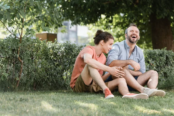 Happy father laughing while sitting near teenage son on green lawn in park — Stock Photo