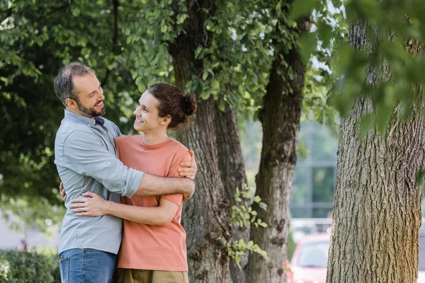 Joyful father with beard hugging happy teenager son in green park — Stock Photo