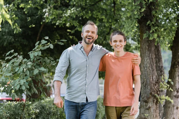 Fröhlicher Vater mit Bart umarmt überglücklichen Teenager-Sohn beim Spaziergang im grünen Park — Stockfoto