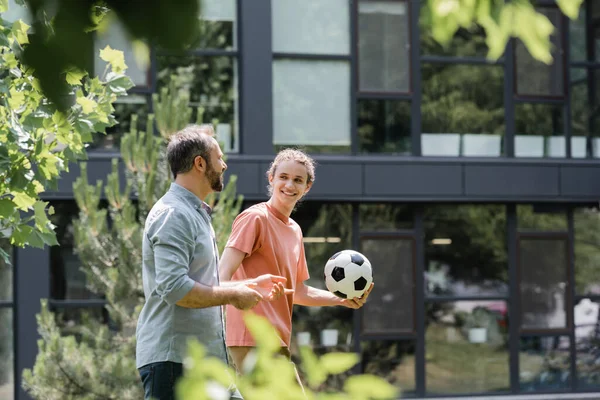 Glücklicher Teenager, der Fußball hält und Vater beim Spaziergang im Freien ansieht — Stockfoto