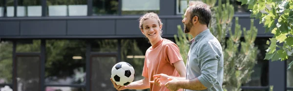Feliz adolescente sosteniendo el fútbol y mirando al padre durante el paseo al aire libre, pancarta - foto de stock