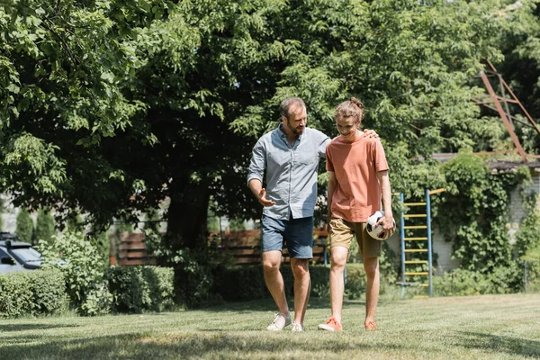 Père barbu étreignant son fils tenant le football tout en marchant dans le champ vert — Photo de stock