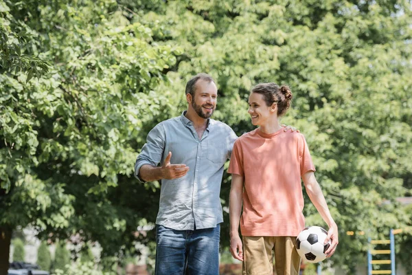 Happy father hugging teenage son holding football while walking in green park — Stock Photo