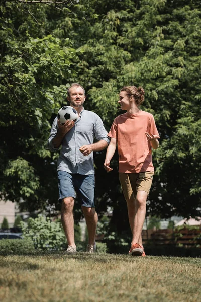 Uomo allegro che tiene il calcio mentre cammina con figlio adolescente in campo verde — Foto stock