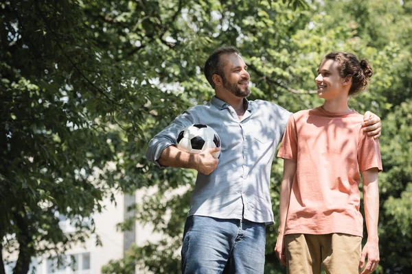 Fröhlicher Mann hält Fußball in der Hand, während er seinen glücklichen Sohn draußen umarmt — Stockfoto