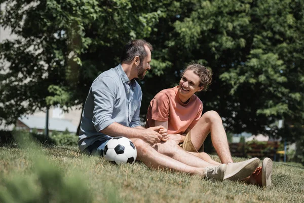 Feliz adolescente sentado cerca de la pelota de fútbol con el padre en el verde parque de verano - foto de stock