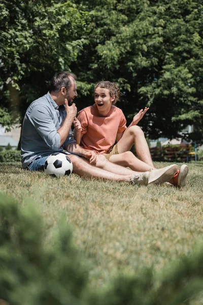 Изумленный подросток, сидящий рядом с футбольным мячом с отцом в зеленом летнем парке — стоковое фото
