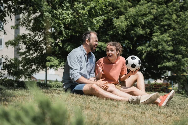 Excited teenage boy sitting near father and soccer ball in green summer park — Stock Photo