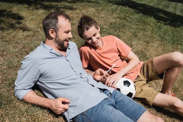 Счастливый отец улыбается во время использования смартфона и отдыхает на лужайке рядом с сыном-подростком после игры в футбол — стоковое фото