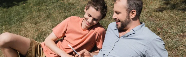 Бородатий батько посміхається під час використання смартфона і відпочиває на газоні біля сина-підлітка, банер — стокове фото