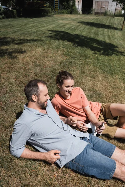 Bearded father smiling while looking at smartphone and resting on lawn near teenage son after playing football — Stock Photo