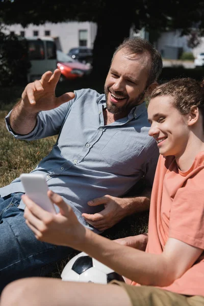 Padre alegre y feliz hijo adolescente sonriendo mientras mira el teléfono inteligente - foto de stock
