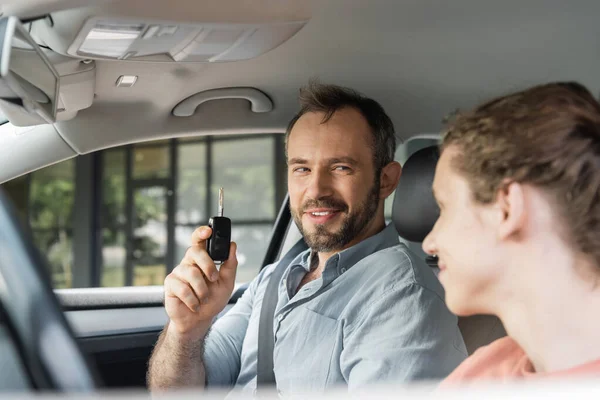 Happy and bearded man holding car key next to son on blurred foreground — Stock Photo