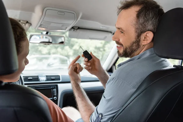Cheerful and bearded dad pointing at car key next to son in modern auto — Stock Photo