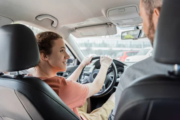 Cheerful teenager holding steering wheel and looking at father in car — Stock Photo