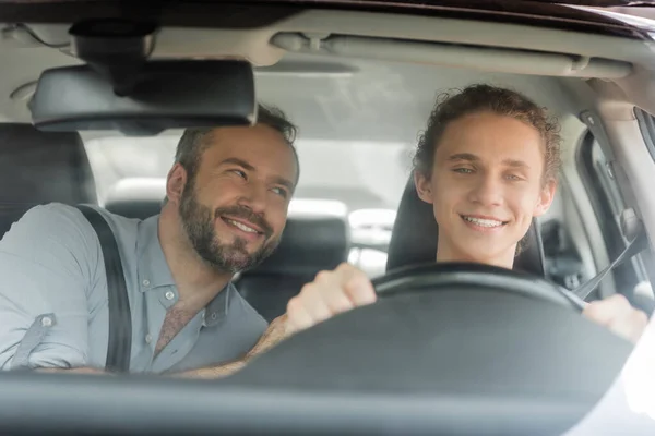 Smiling teenager boy holding steering wheel while driving car next to dad — Stock Photo