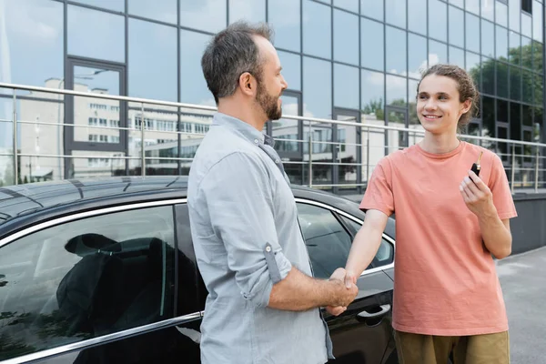 Happy bearded man shaking hands with cheerful teenage son holding car key near automobile — Stock Photo