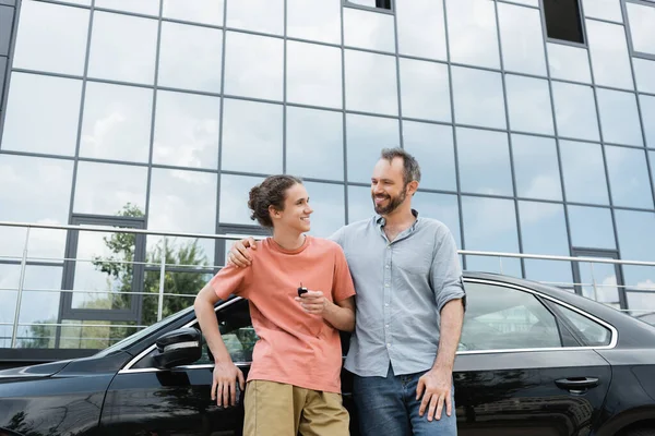 Счастливый бородатый мужчина кладет руку на плечо веселого сына-подростка с ключом от машины рядом с автомобилем — стоковое фото