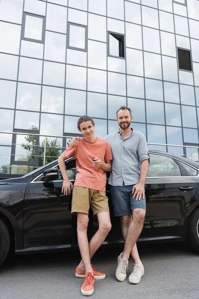 Happy bearded father putting hand on shoulder of cheerful teenage son holding car key near automobile — Stock Photo