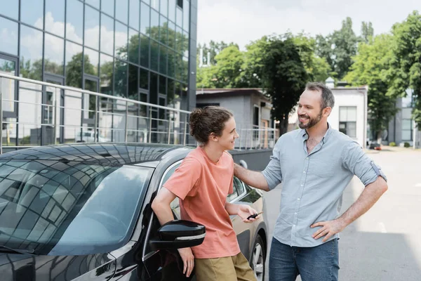 Счастливый бородатый отец смотрит на веселого сына-подростка с ключом от машины рядом с автомобилем — стоковое фото