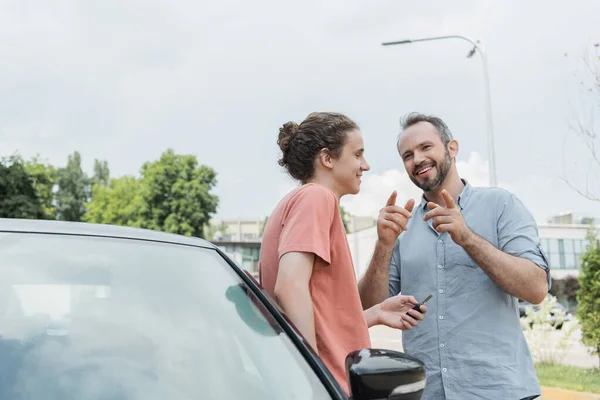 Щасливий бородатий тато вказує пальцями біля сина-підлітка, що стоїть біля машини з ключами — стокове фото