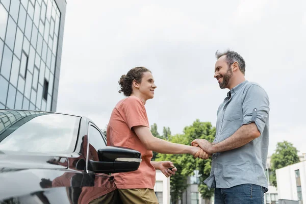 Low angle view of happy father and teenage son holding hands near modern car — Stock Photo