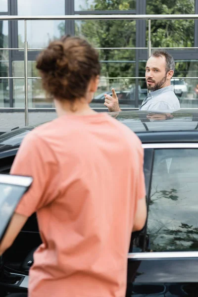 Бородатий батько вказує на сина-підлітка, стоячи разом біля сучасної машини — стокове фото