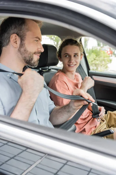 Happy teenage boy and bearded dad smiling while fastening safety belts in car — Stock Photo