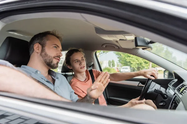 Бородатий тато вказує, показуючи напрямок до підліткового сина за кермом автомобіля — стокове фото
