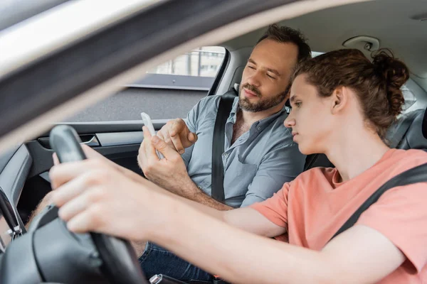 Pai barbudo apontando para smartphone enquanto sentado ao lado de filho adolescente no carro — Fotografia de Stock