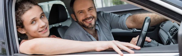 Happy father and smiling teenage son looking at camera while sitting together in car, banner — Stock Photo