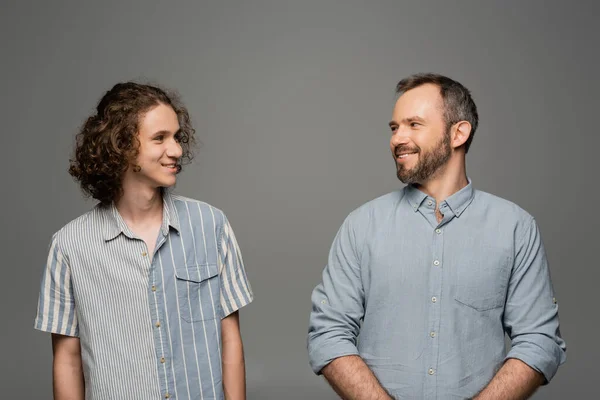 Cheerful and bearded father looking at curly happy son isolated on grey — Stock Photo