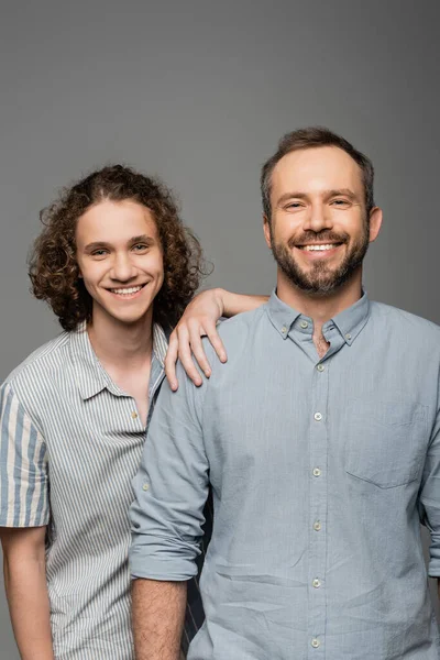 Счастливый отец и сын в рубашках, смотрящих в камеру и улыбающихся изолированных на сером — стоковое фото