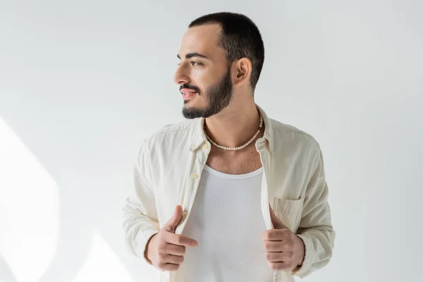 Sonriente y barbudo hombre gay en ropa casual y collar de perlas tocando camisa y mirando hacia otro lado mientras posando sobre fondo gris con luz solar - foto de stock
