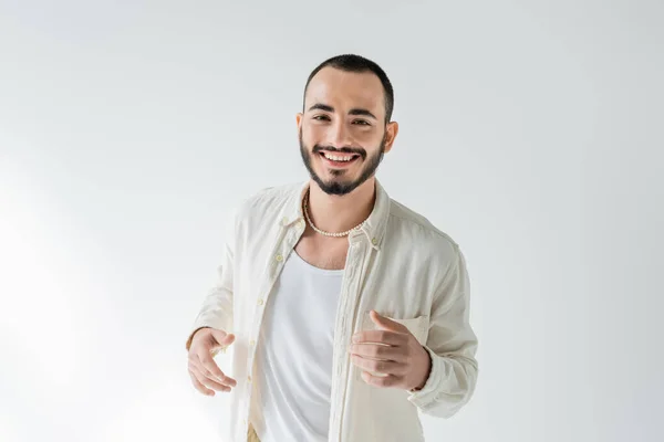 Cheerful and bearded young homosexual man in beige shirt and pearl necklace looking at camera while posing on grey background — Stock Photo