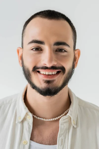 Ritratto di giovane e felice uomo omosessuale in abiti casual e collana di perle guardando la macchina fotografica mentre in piedi e sorridente isolato sul grigio — Foto stock