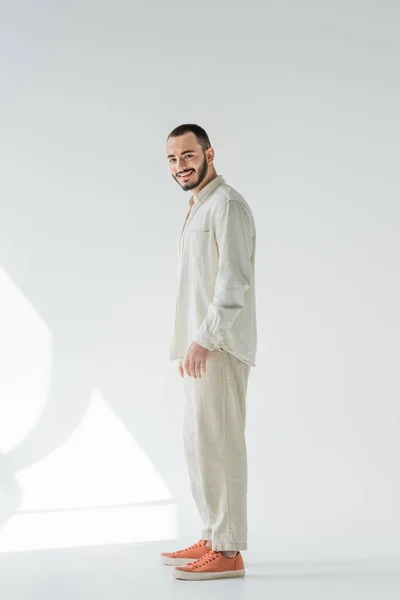 Longitud completa de hombre homosexual sonriente y barbudo en ropa beige con telas naturales mirando a la cámara mientras está de pie sobre fondo gris con luz solar - foto de stock