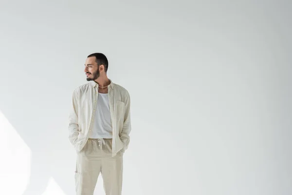 Smiling homosexual man in beige clothes and pearl necklace holding hands in pockets of pants and looking away while standing on grey background with sunlight — Stock Photo