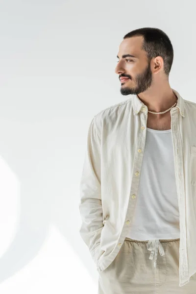 Bearded and short haired homosexual man in beige shirt and stylish pearl necklace looking away and holding hand in pocket of pants made of natural fabrics on grey background with sunlight — Stock Photo