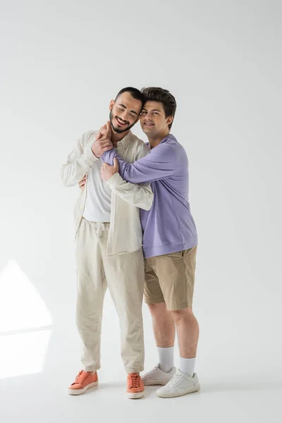 Full length of smiling gay man in casual sweatshirt and shorts hugging bearded boyfriend with closed eyes on grey background with sunlight — Stock Photo