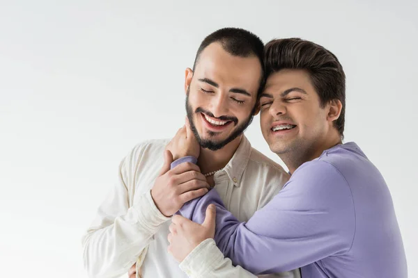Беззаботный молодой гей в брекетах в повседневной одежде обнимает и трогает шею брюнетки и бородатого партнера с закрытыми глазами, изолированными на сером — стоковое фото