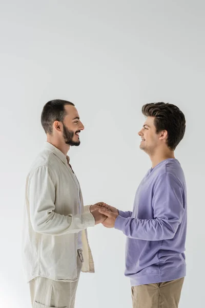 Вид сбоку улыбающейся гомосексуальной пары в повседневной одежде, смотрящей друг на друга и держащейся за руки, находясь в изоляции на сером — стоковое фото