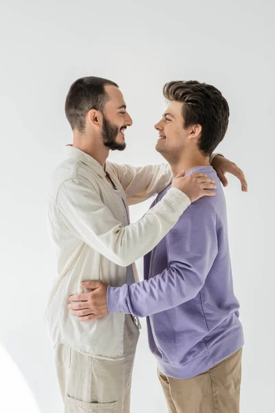 Vista lateral de pareja homosexual joven y alegre en ropa casual abrazándose y mirándose mientras están de pie sobre un fondo gris - foto de stock